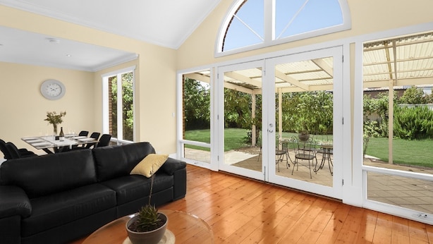 A spacious living room with a black sofa, a dining table, and a large glass door leading to an outdoor patio with a garden view. - Adelaide Style Accommodation-City-Beach (Everard Park)