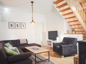 Una sala de estar moderna con un sofá, un sillón y un televisor sobre un mueble. Se ve una escalera de madera al fondo.