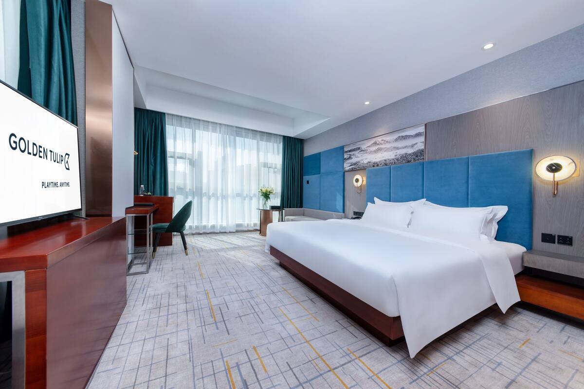 A hotel room with a large bed, a desk with a chair, a TV, and a wall-mounted lamp.