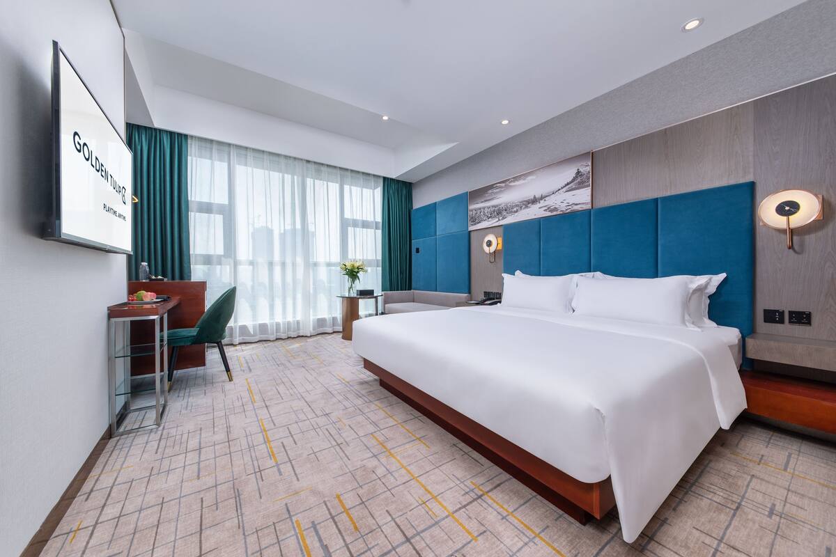 A modern hotel room with a large bed, a desk with a chair, a TV, and a view of the outdoors.