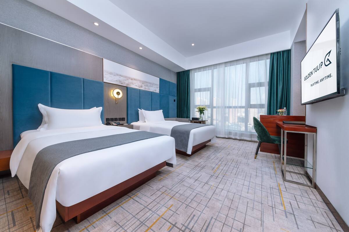 A hotel room with two beds, a desk, a chair, a TV, and a large window with curtains.