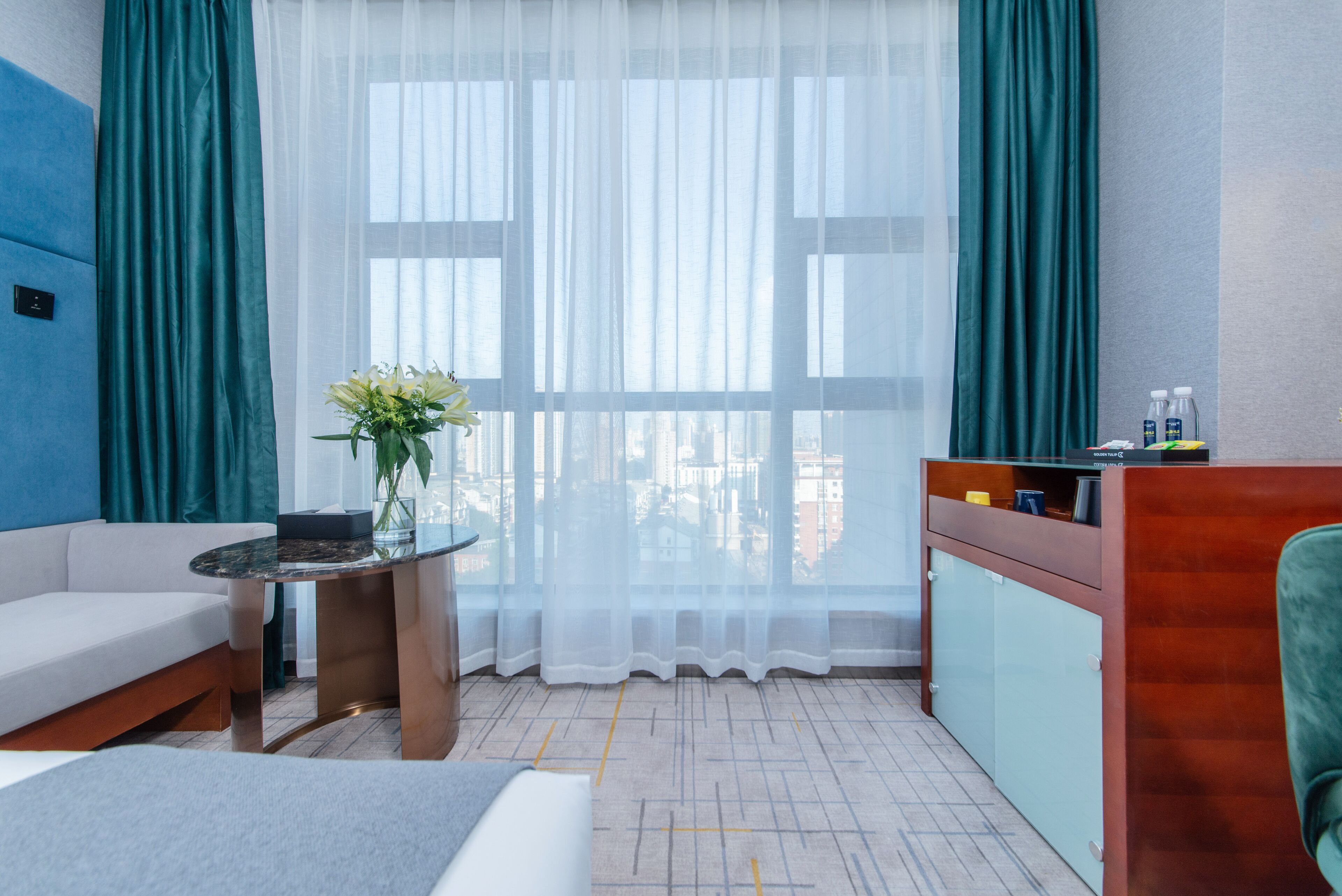 A modern hotel room with a sofa, a small round table, a desk with a chair, and a view of the city through sheer curtains.
