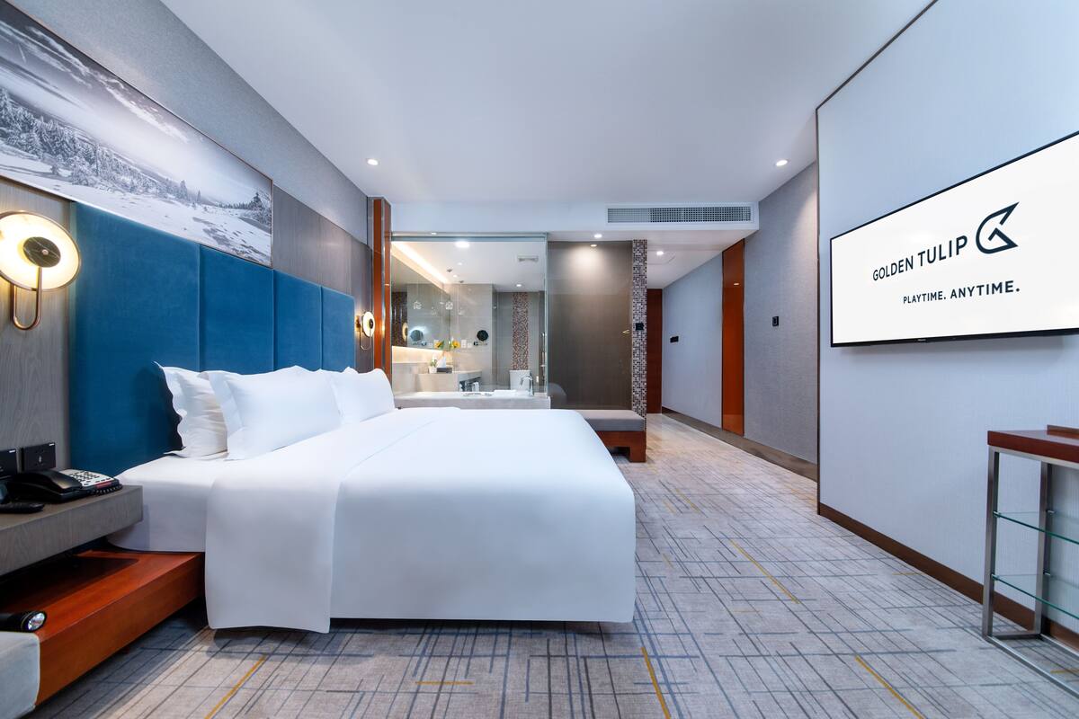 A modern hotel room with a large bed, a desk with a phone, a mirror, and a wall-mounted TV displaying the hotel's name.