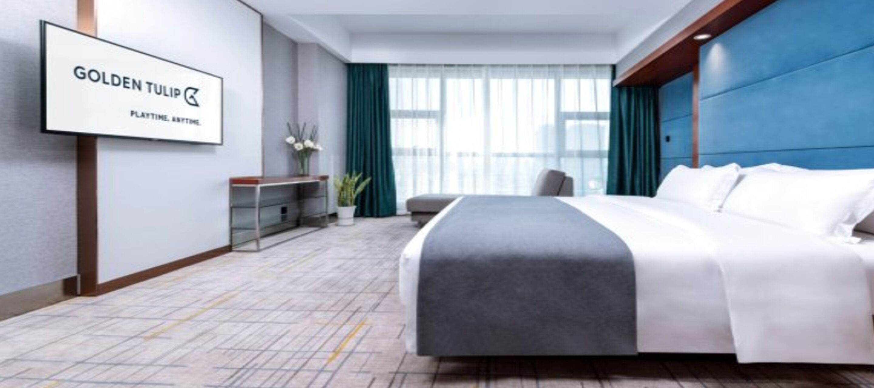 A hotel room with a bed, a desk, and a chair. There is a window with curtains and a sign on the wall.