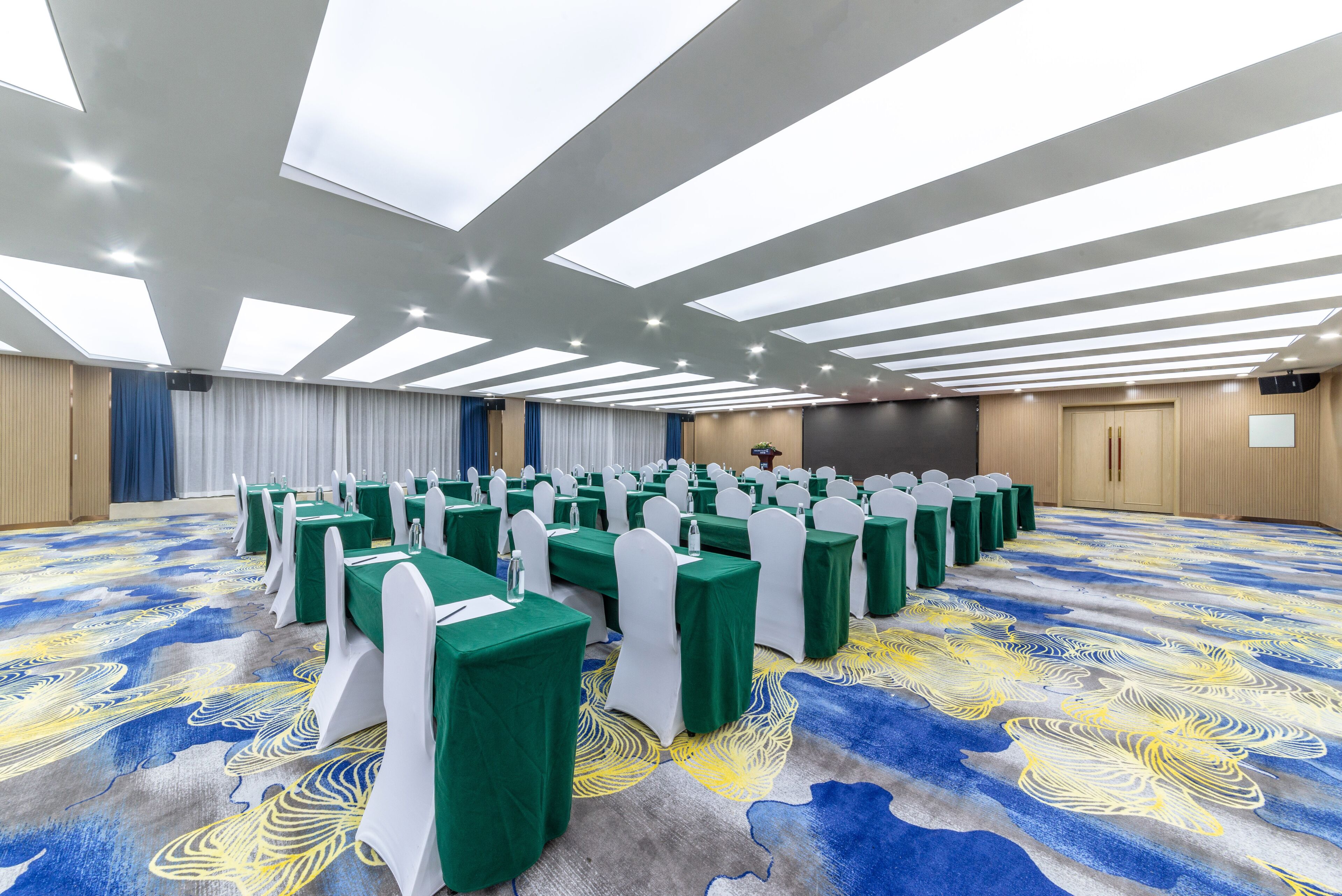 Un salón de conferencias con filas de mesas, sillas blancas y una alfombra estampada.