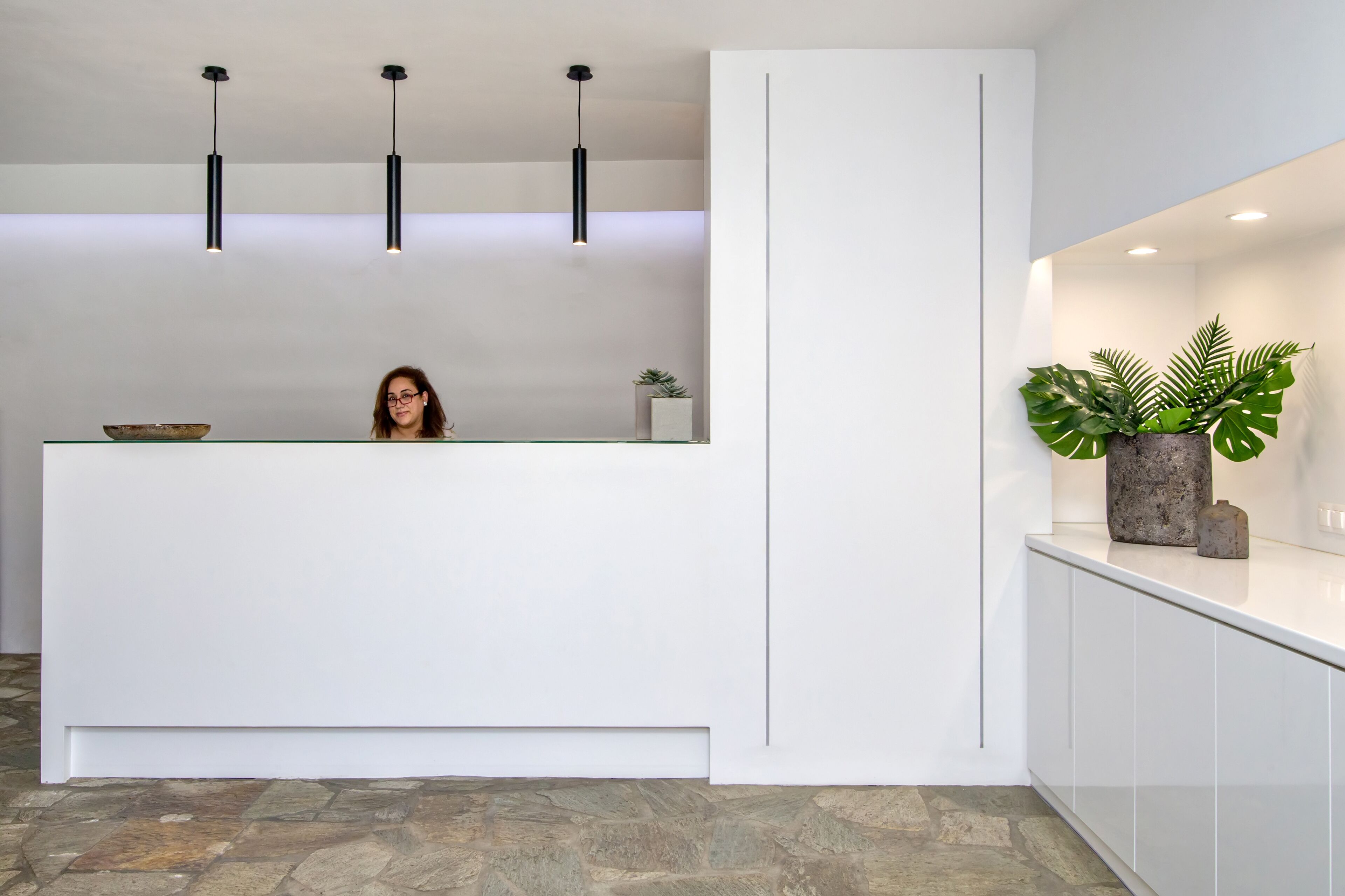 Un espace d’accueil avec un comptoir de réception blanc, des luminaires suspendus, une plante en pot et un sol carrelé en pierre.
