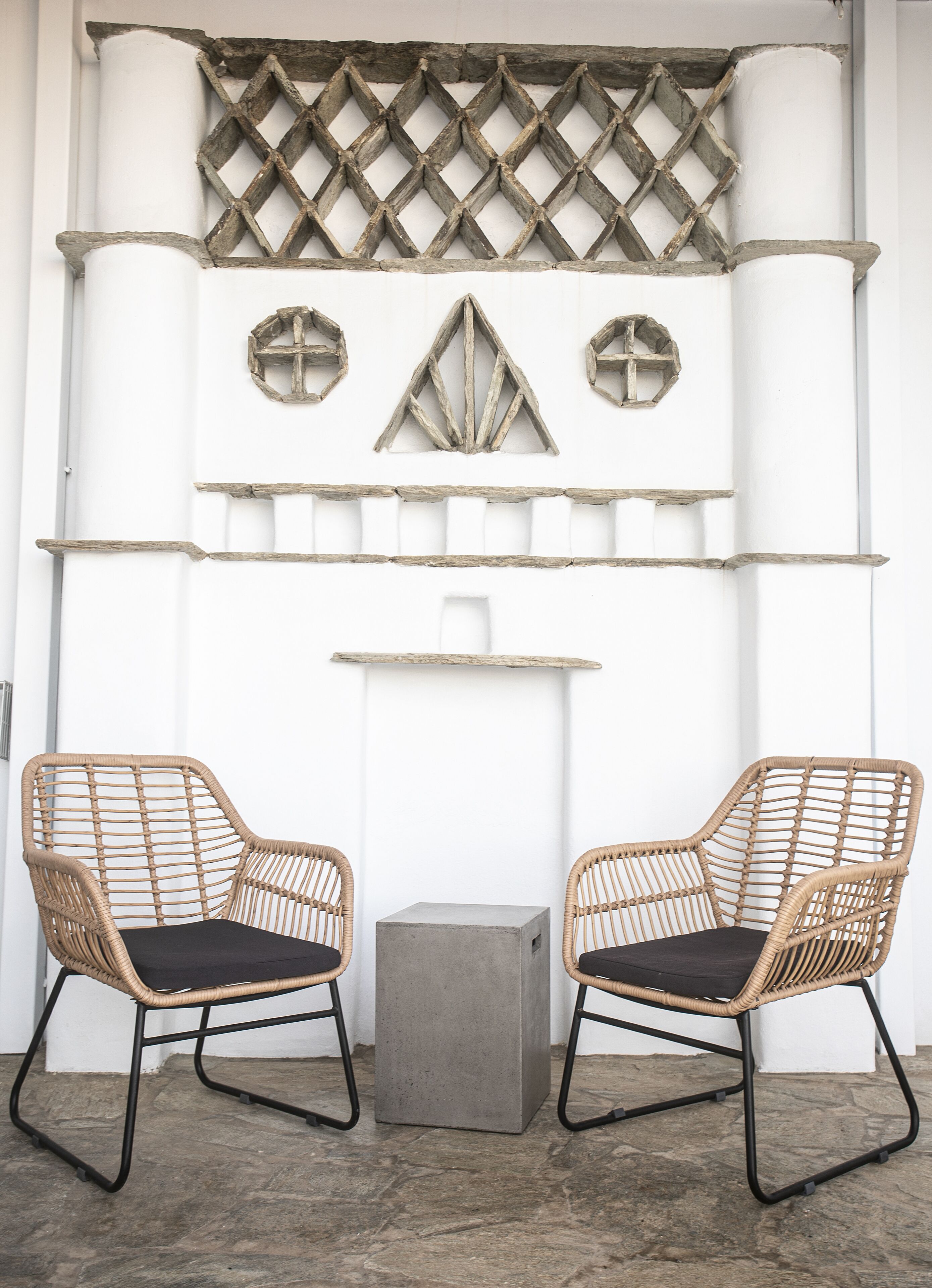 Deux fauteuils en osier avec des coussins noirs, une petite table en béton et un mur décoratif orn é de motifs géométriques et d’éléments en bois.