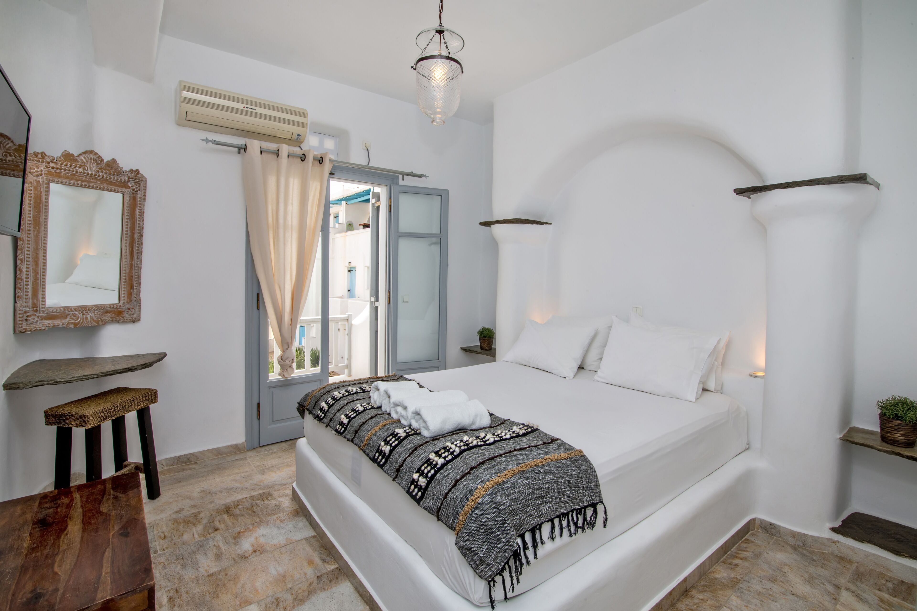 A bedroom with a bed, a mirror, a window with curtains, a small table, and a wall-mounted shelf.