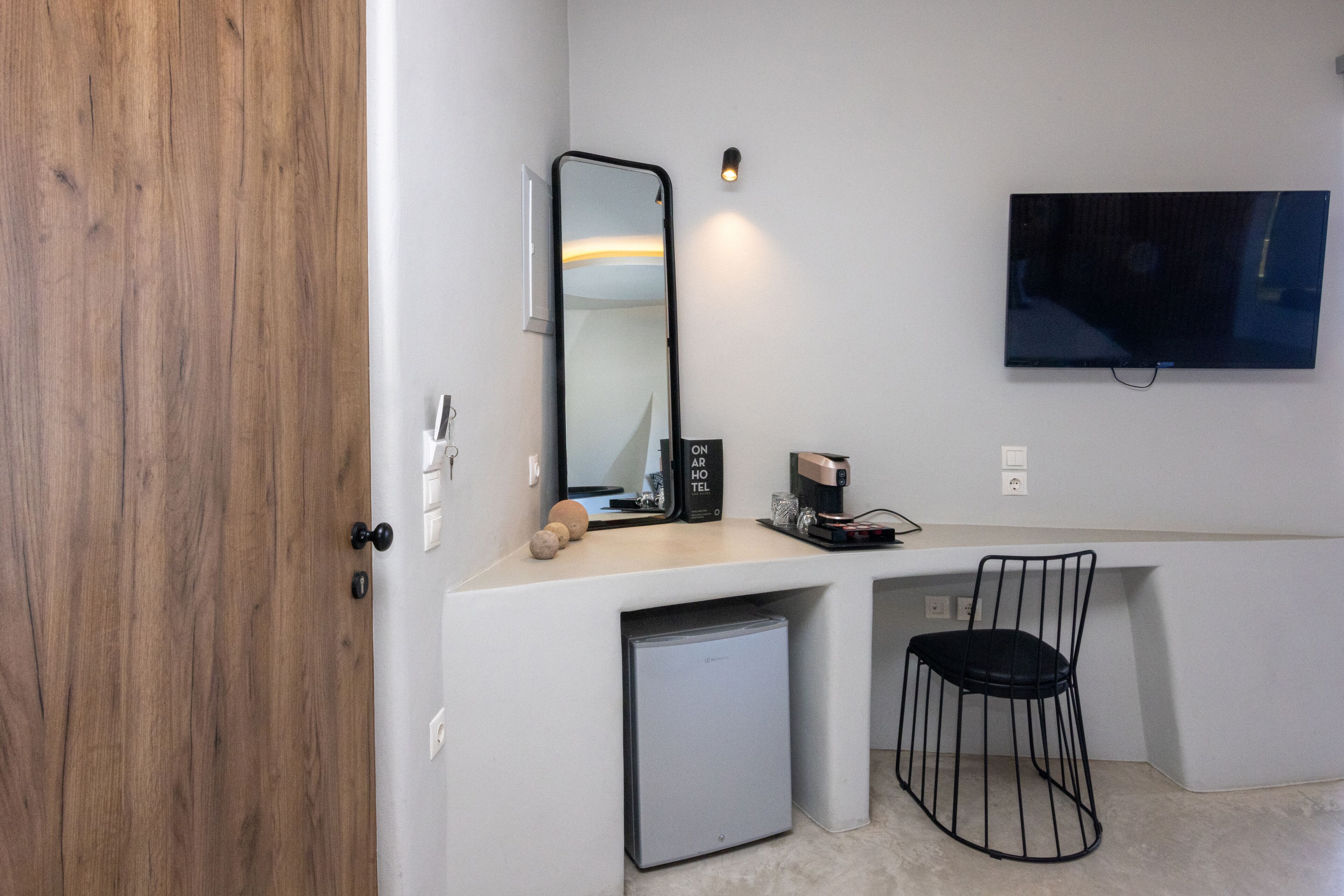 A compact room with a wooden door, a small kitchenette, a mirror, a TV, and a black chair.