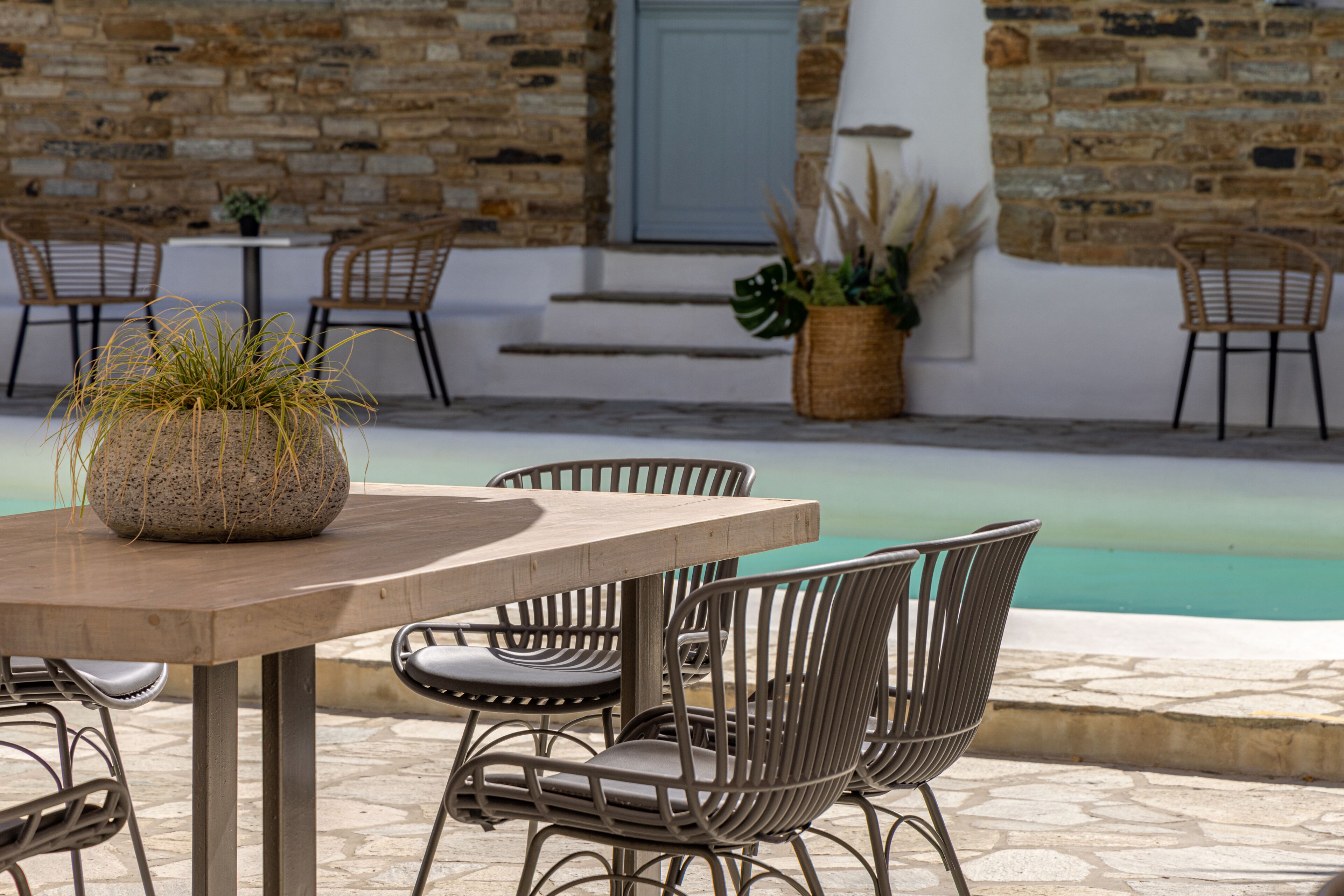 An outdoor seating area with a round table and metal chairs, a stone wall, and a pool in the background.