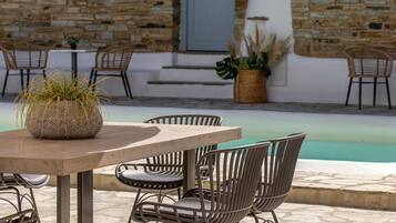 An outdoor seating area with a round table and metal chairs, a stone wall, and a pool in the background.