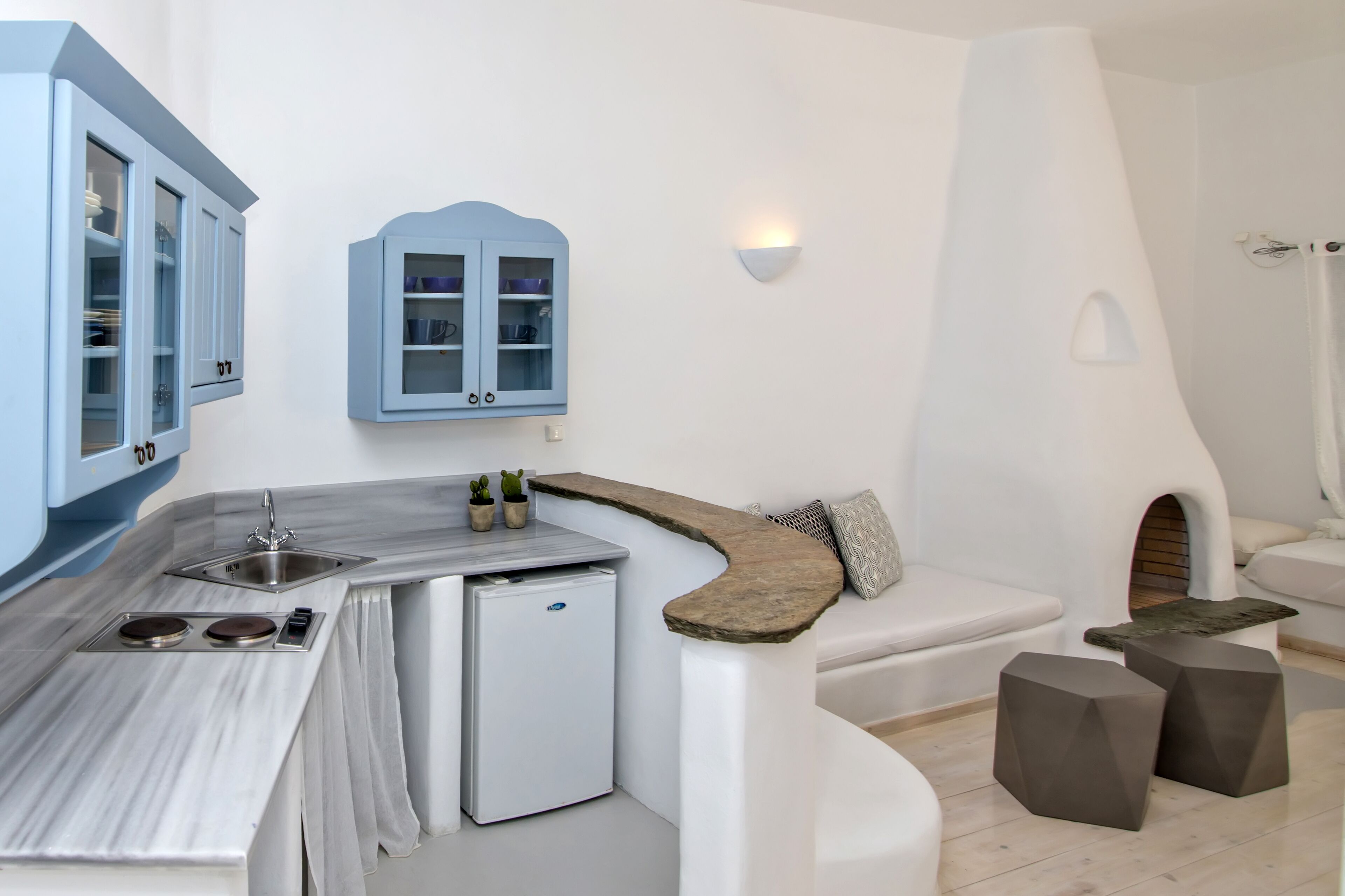 A compact kitchen with a built-in refrigerator, a sink, and a small countertop. There is a white sofa with a cushion and two dark stools.