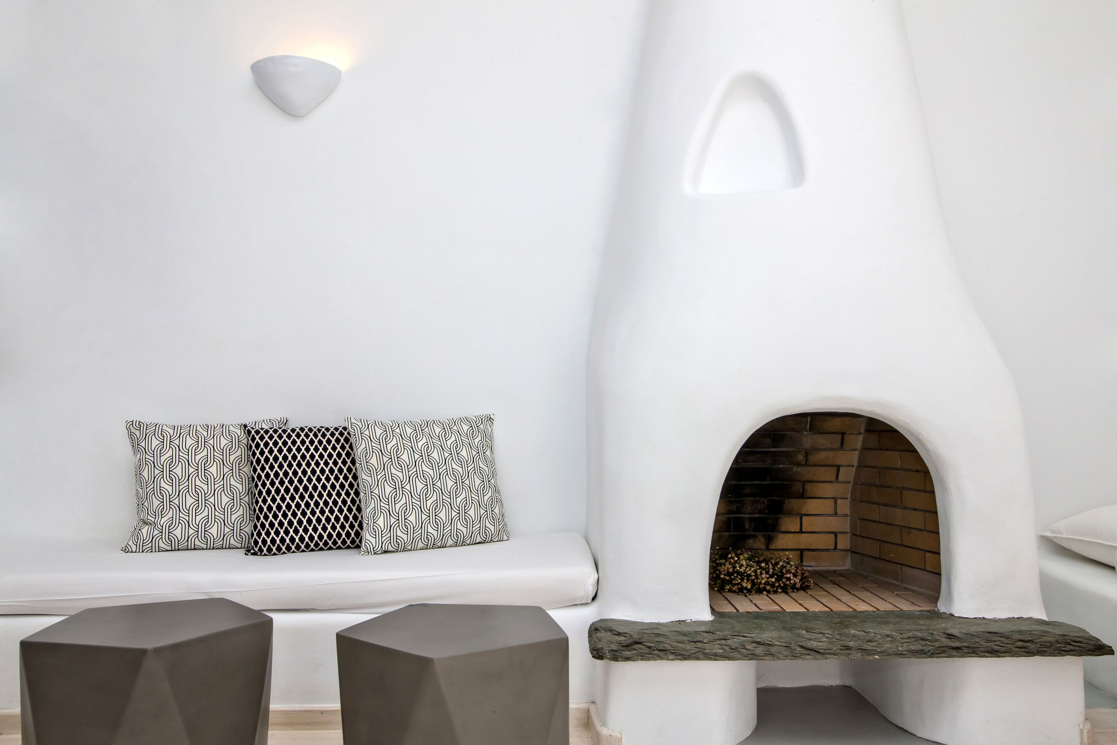A modern interior with a white wall, a built-in fireplace, a white sofa with patterned cushions, and two dark stools.