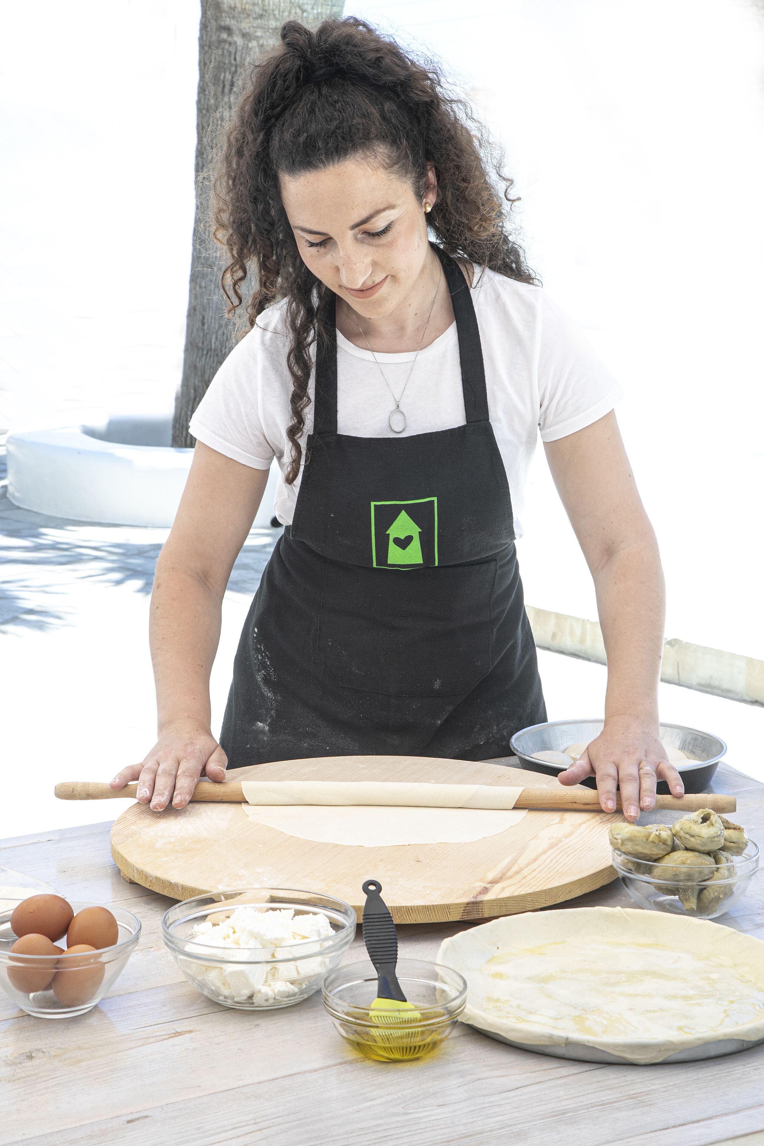 A person rolling dough on a wooden board with a rolling pin, surrounded by bowls of ingredients like eggs, cheese, and oil.