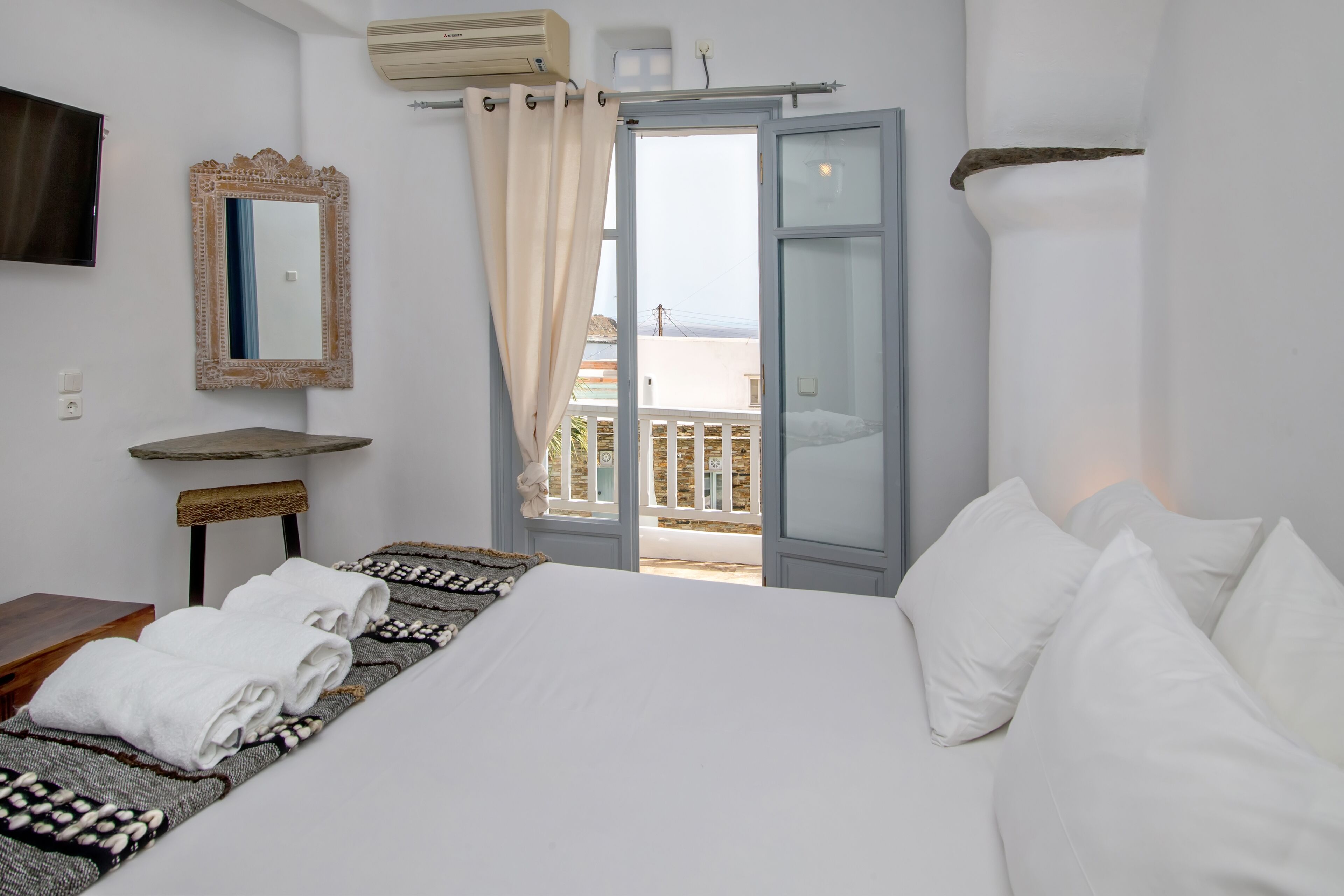 A hotel room with a bed, a small table, a mirror, and a view of the outside balcony.