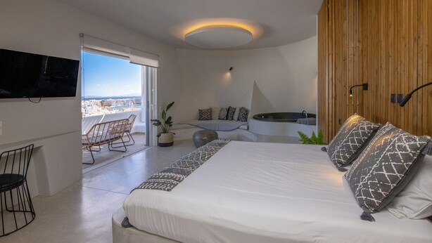 Ein modernes Schlafzimmer mit einem großen Bett, einem an der Wand befestigten Fernseher, einer Sitzecke mit Sesseln und Blick auf die Stadt durch große Fenster.