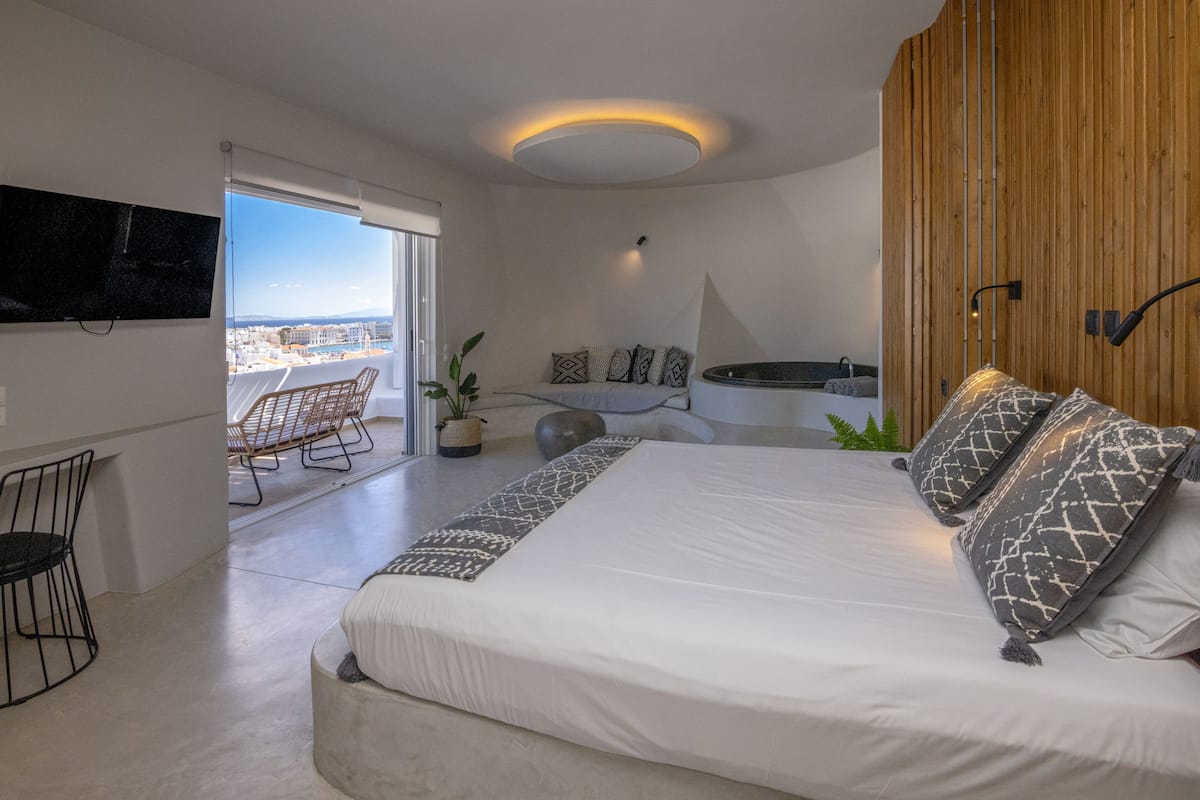 Um quarto moderno com uma cama grande, uma televisão na parede, um espaço de estar com cadeiras e vista da cidade por grandes janelas.