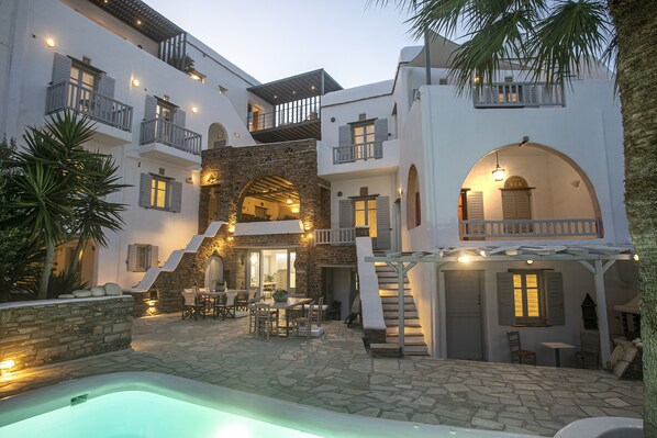A multi-story building with a stone courtyard, outdoor seating area, and a swimming pool. - Onar Hotel and Suites (Tinos)
