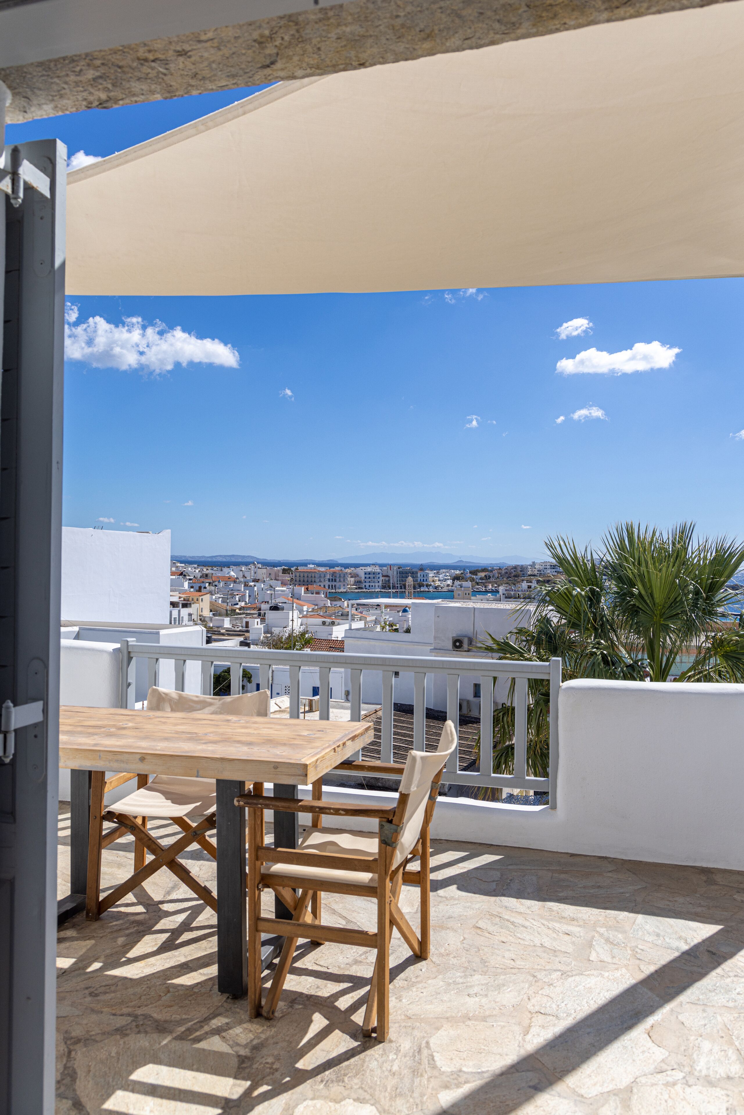 A terrace with a wooden table and chairs, a view of a coastal city, and a clear sky.
