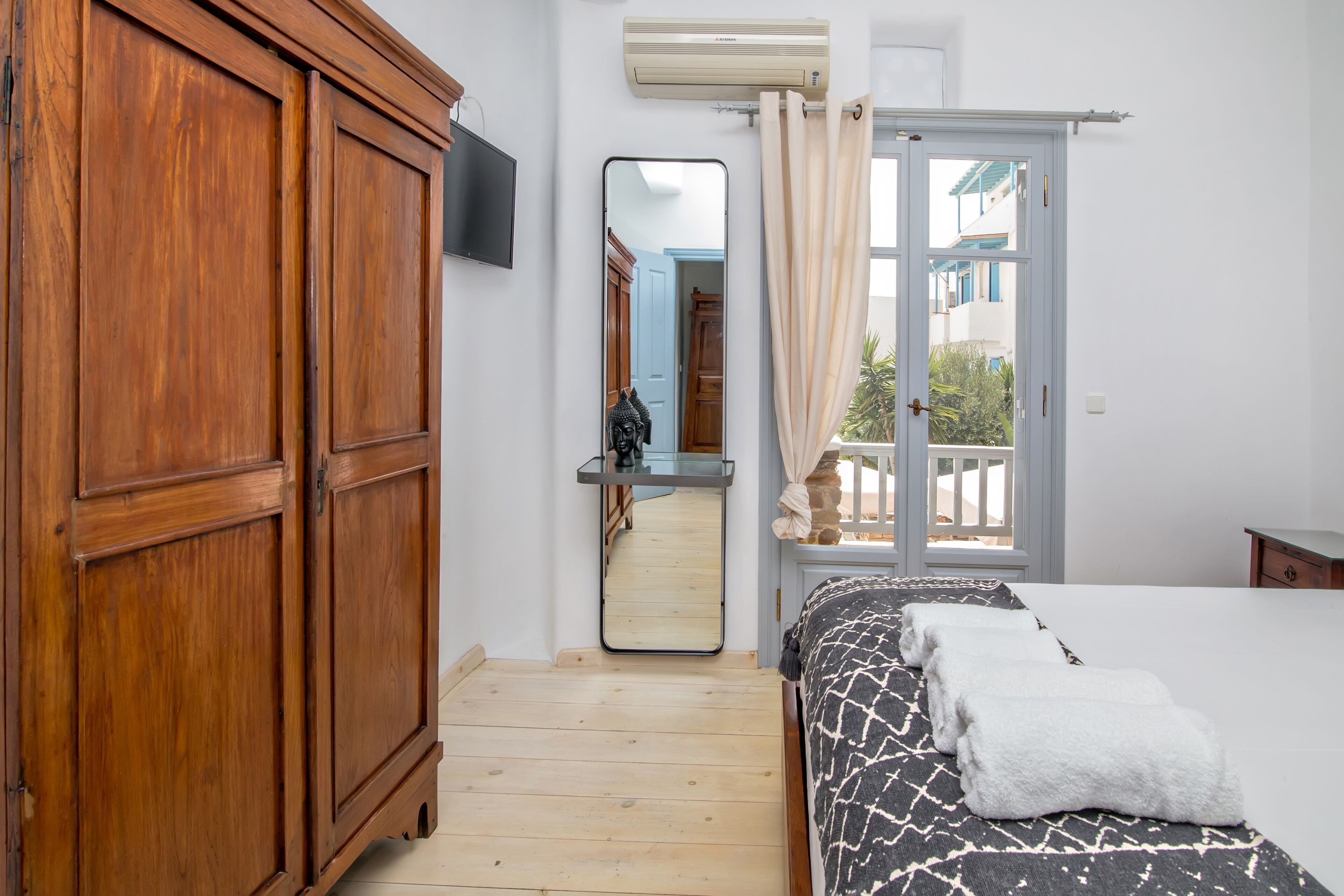 A bedroom with a wooden wardrobe, a bed with a patterned bedspread, a mirror, and a window with curtains.