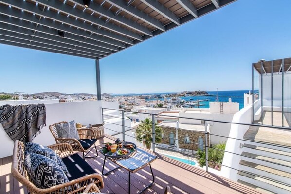 A balcony with wicker furniture, a table with a drink and snacks, and a view of the sea and cityscape. - Onar Hotel and Suites (Tinos)