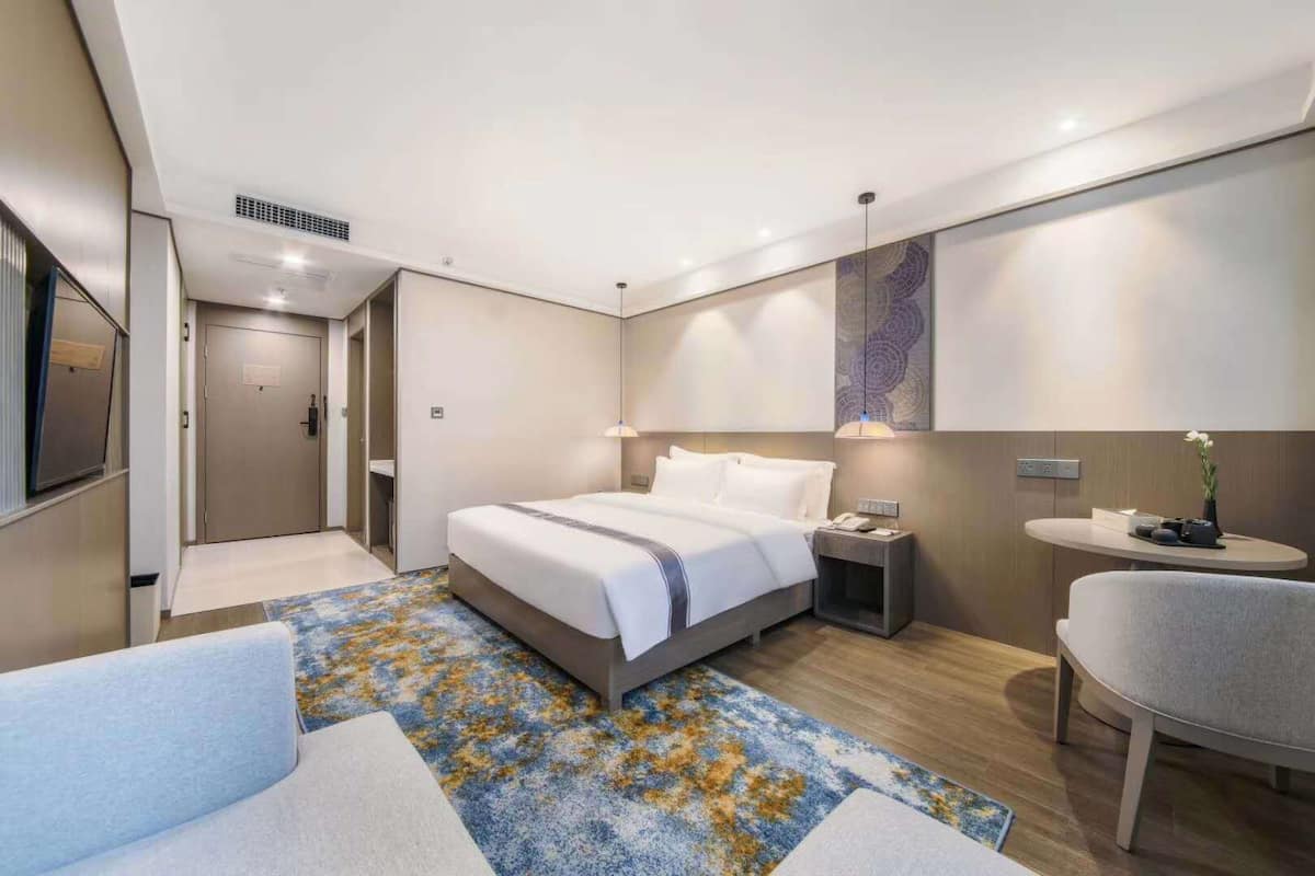 Una habitación de hotel moderna con una cama grande, mesita de noche, una silla y un escritorio pequeño con lámpara.