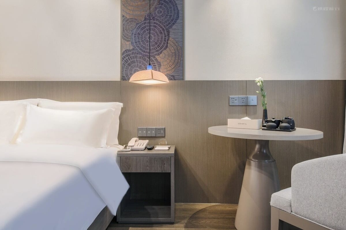 Habitación de hotel con cama, mesita de noche, lámpara y una pequeña mesa redonda con un florero.
