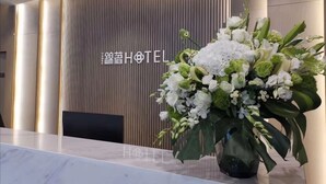 A hotel reception desk with a modern design, featuring a vertical wooden panel wall and a large floral arrangement in a black vase.