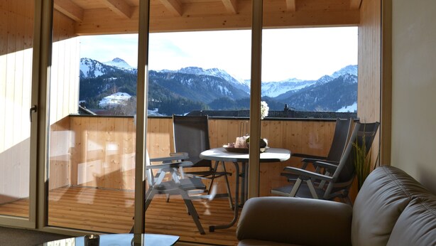 A wooden balcony with a view of snow-capped mountains, a table with chairs, and a sofa. - Dorner Hof (Sibratsgfaell)