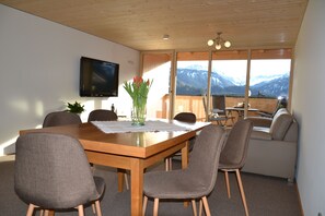 Un comedor con una mesa de madera, sillas grises tapizadas, un jarrón con flores y un televisor montado en la pared.