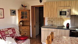 Una cocina acogedora con armarios de madera, un microondas y una estufa. Hay una mesa de comedor con mantel y sillas. Se aprecia un mueble de madera con puertas de cristal, y una puerta conduce a otra habitación.