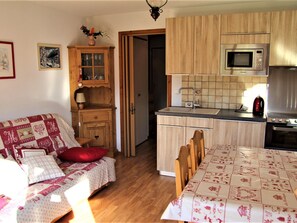 A cozy kitchen with wooden cabinets, a microwave, and a stove. There's a dining table with a tablecloth and chairs. A wooden cabinet with glass doors is visible, and a door leads to another room.