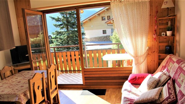 Un cuarto acogedor con piso de madera, un sofá con cojines estampados, una mesa de comedor con sillas y un ventanal corredizo que da a una terraza exterior.
