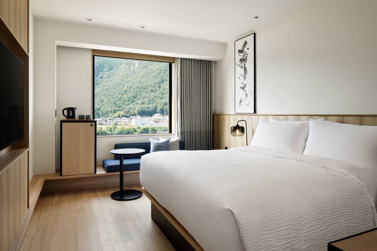 A modern hotel room with a large bed, a small seating area, a view of a forested area, and a flat-screen TV.