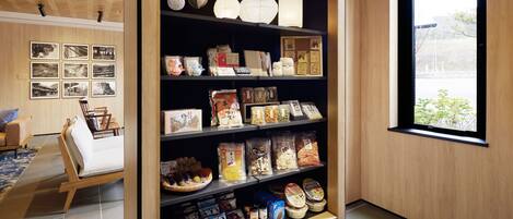 A room with a wooden floor, a sofa, a bookshelf filled with snacks and books, and a window with a view of greenery.