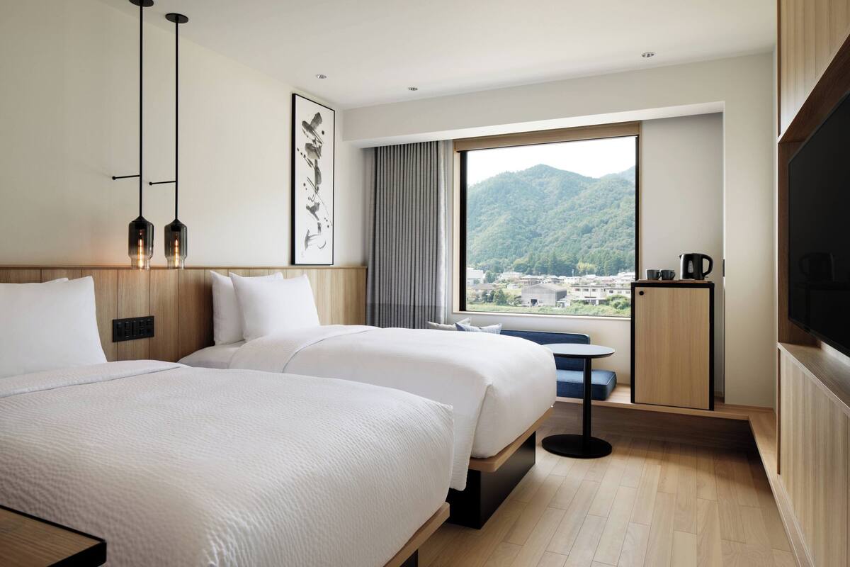 A hotel room with two beds, a large window with a city and mountain view, a small table with a chair, and a television on a wooden cabinet.
