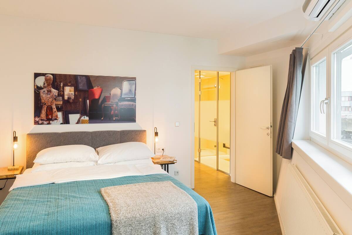 A modern hotel room with a large bed, a bedside table with a lamp, a painting on the wall, and a view of the bathroom through an open door.
