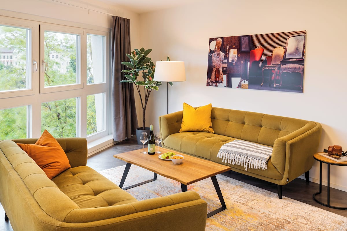 A living room with a sofa, armchair, and coffee table. There is a window with curtains, a lamp, and a painting on the wall.