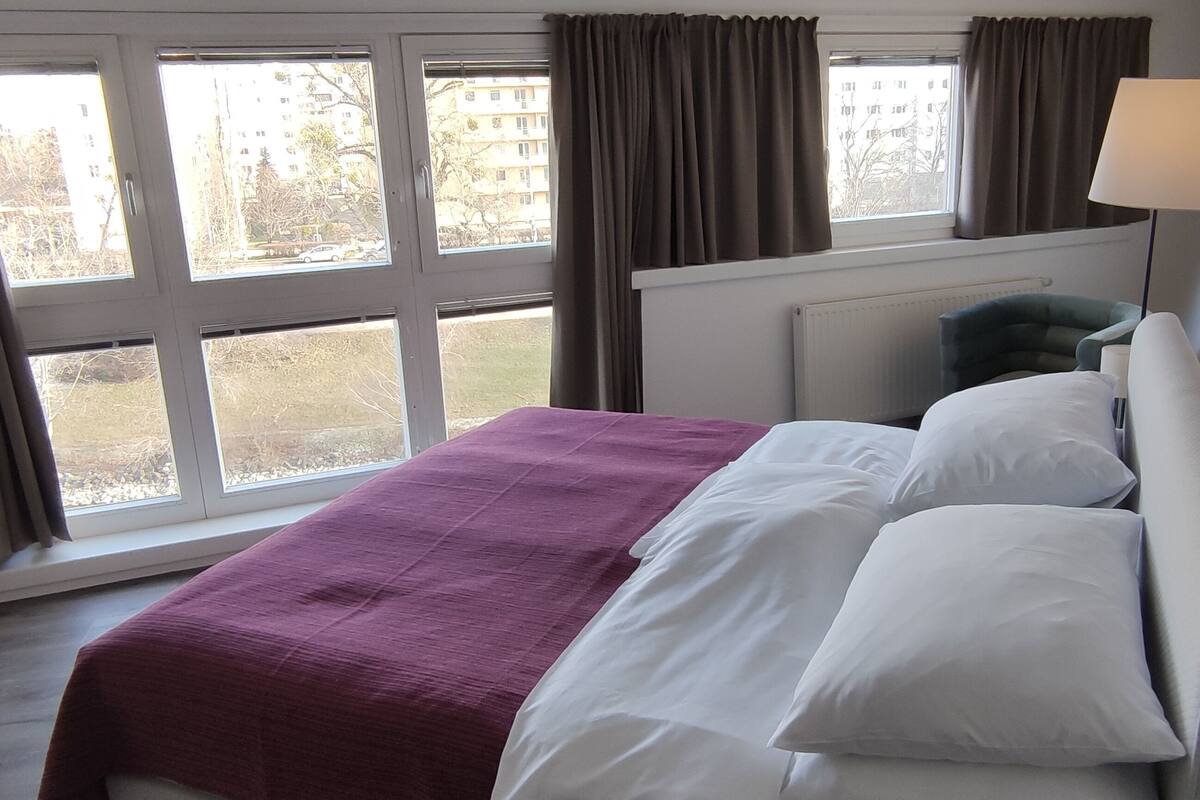A hotel room with a bed, a window with curtains, a lamp, and a view of buildings outside.