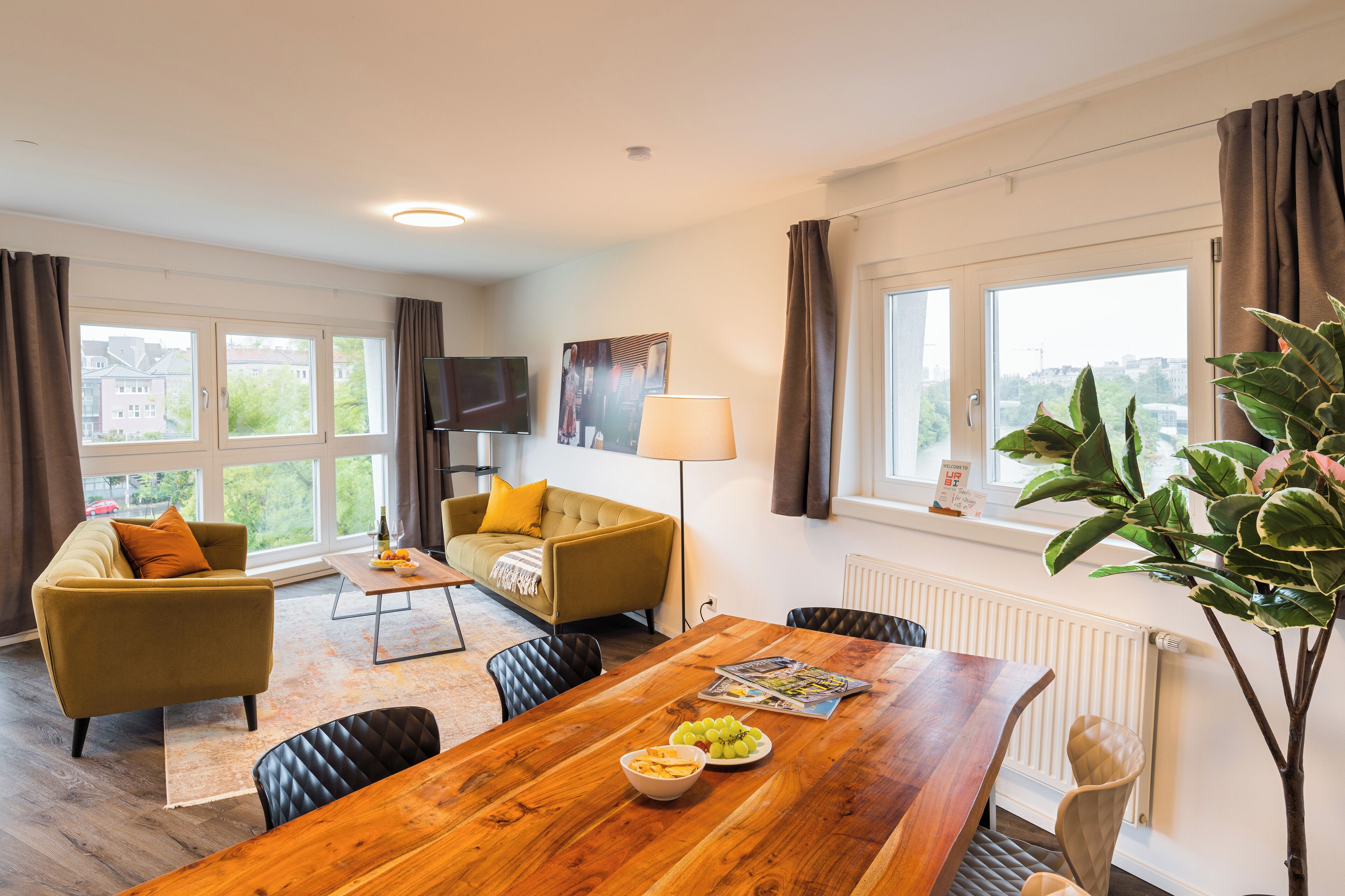 Un salon moderne avec un canapé, une table à manger ornée de fruits, une lampe et une vue sur la ville grâce à de grandes fenêtres.