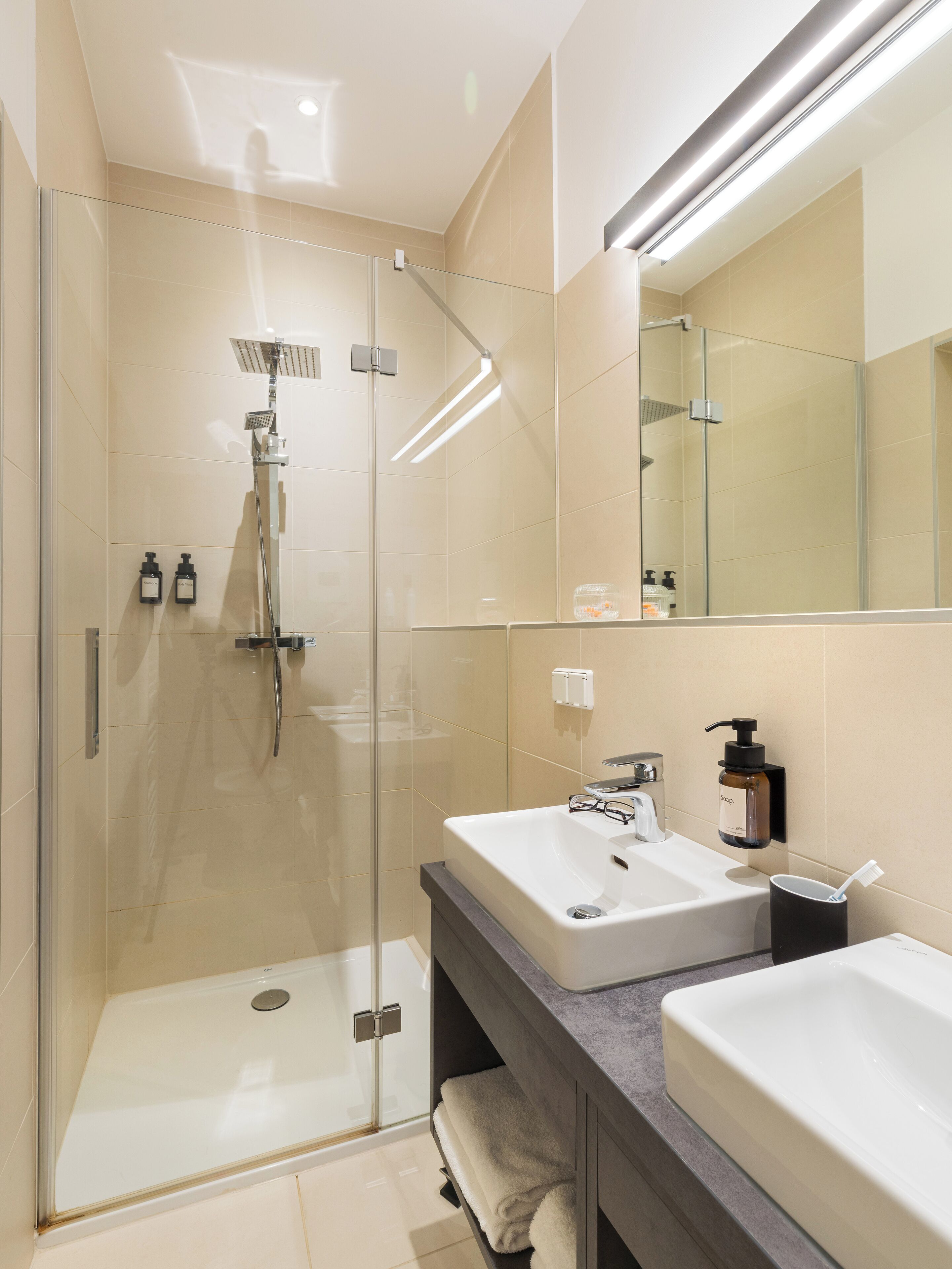 Un baño moderno con ducha de vidrio, dos lavamanos y un espejo grande.