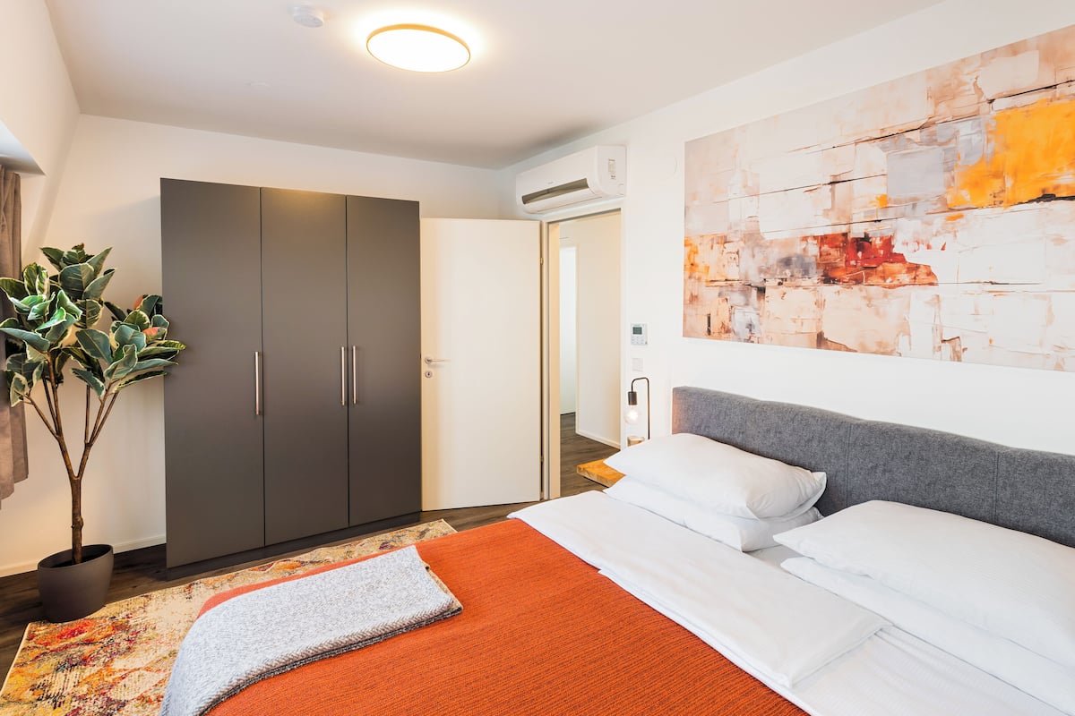 A modern hotel room with a large bed, a grey wardrobe, a potted plant, and a colorful abstract painting on the wall.