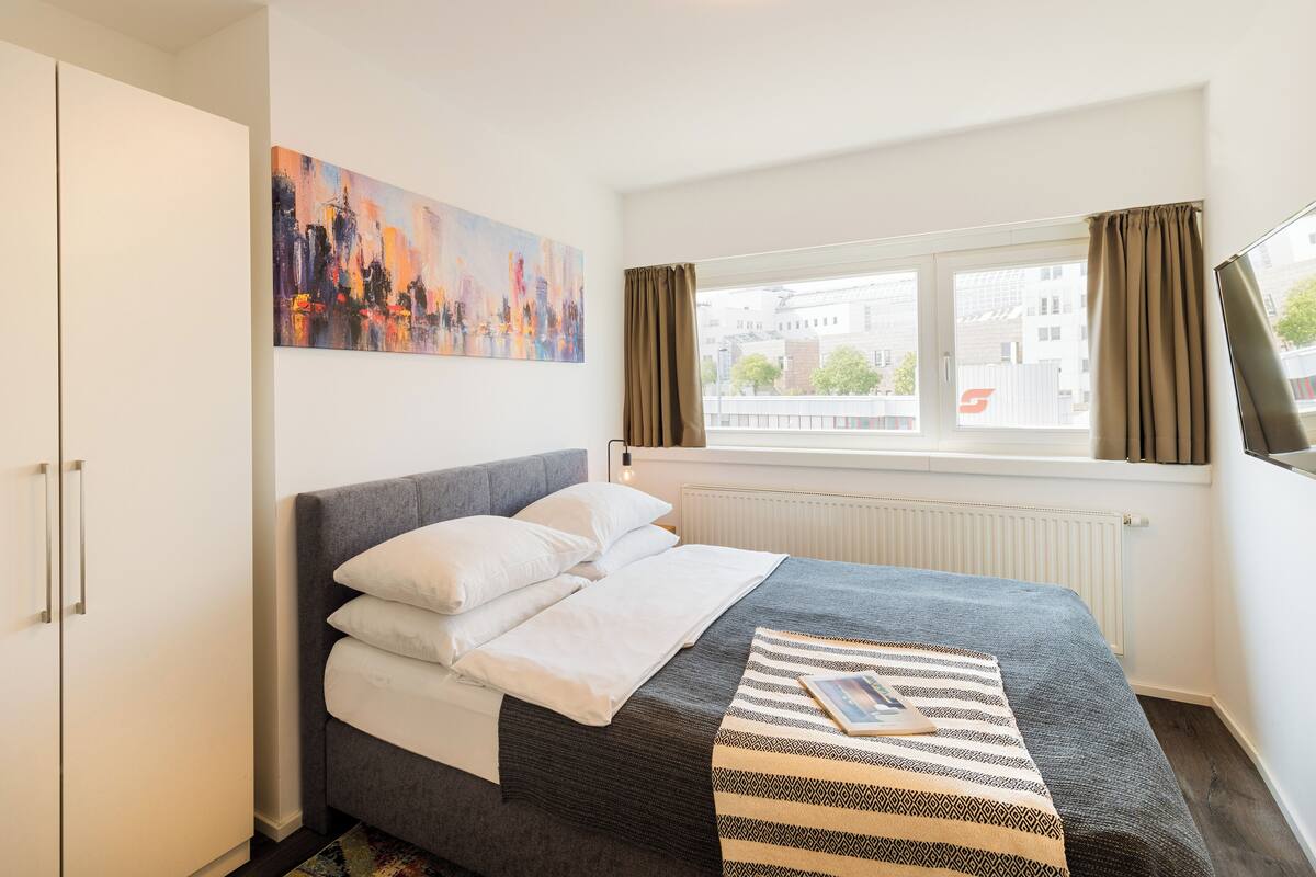 A neatly arranged bedroom with a bed, a painting, a window with curtains, and a radiator.