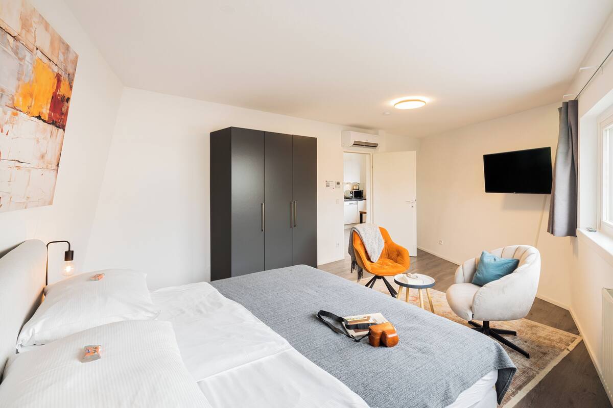 A modern hotel room with a bed, a desk, an orange chair, a grey sofa, and a flat-screen TV.