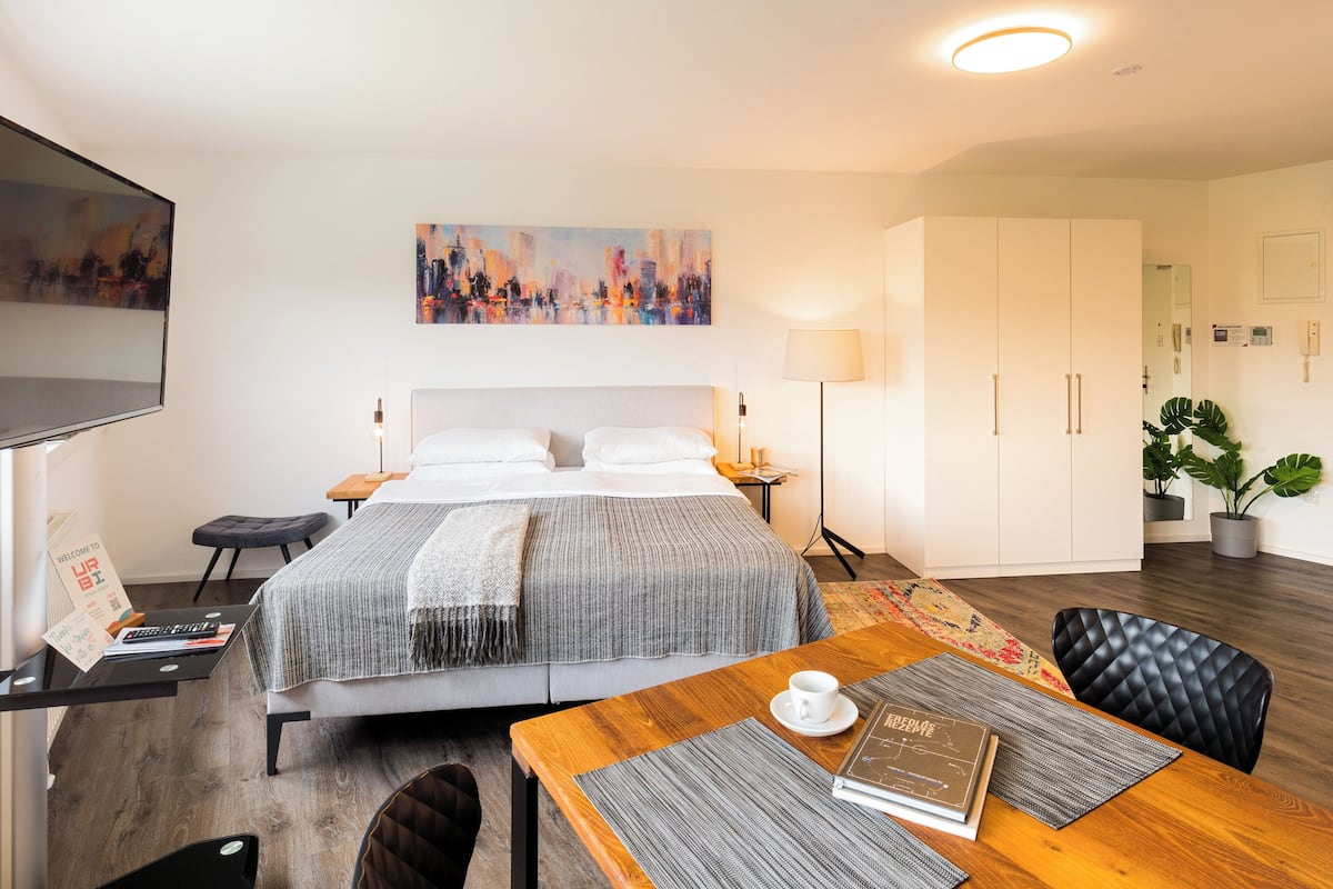 A modern hotel room with a bed, a desk with a laptop, a coffee cup, and a plant.