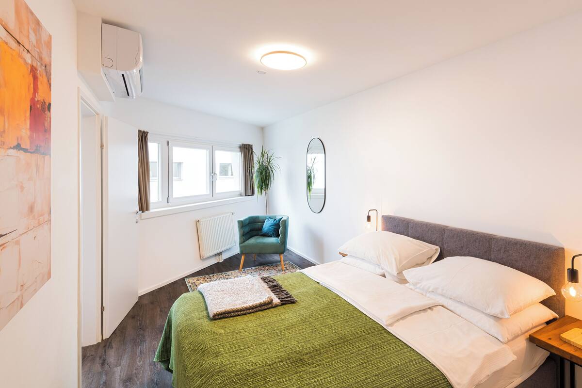 A bedroom with a bed, a green throw, a window, a plant, and a mirror.