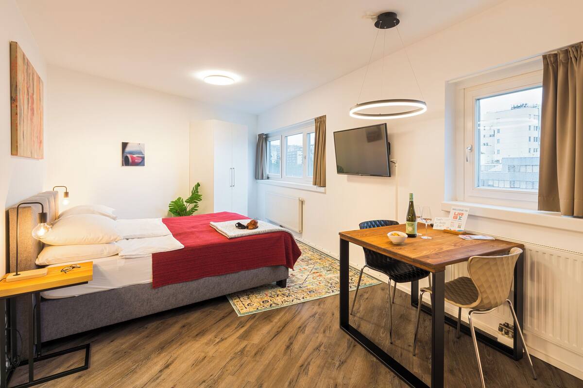 A modern hotel room with a bed, a desk with a chair, a TV, and a window with curtains.