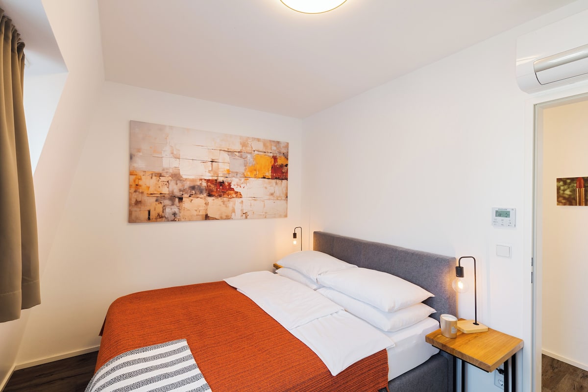 A neatly made bed with white and orange bedding, a modern abstract painting on the wall, and a bedside table with a lamp.
