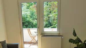 A modern living room with a grey sofa, two round tables, a large window with a view of greenery, and a radiator. - Glück Auf Appartements Walter-Hohmann (Essen)
