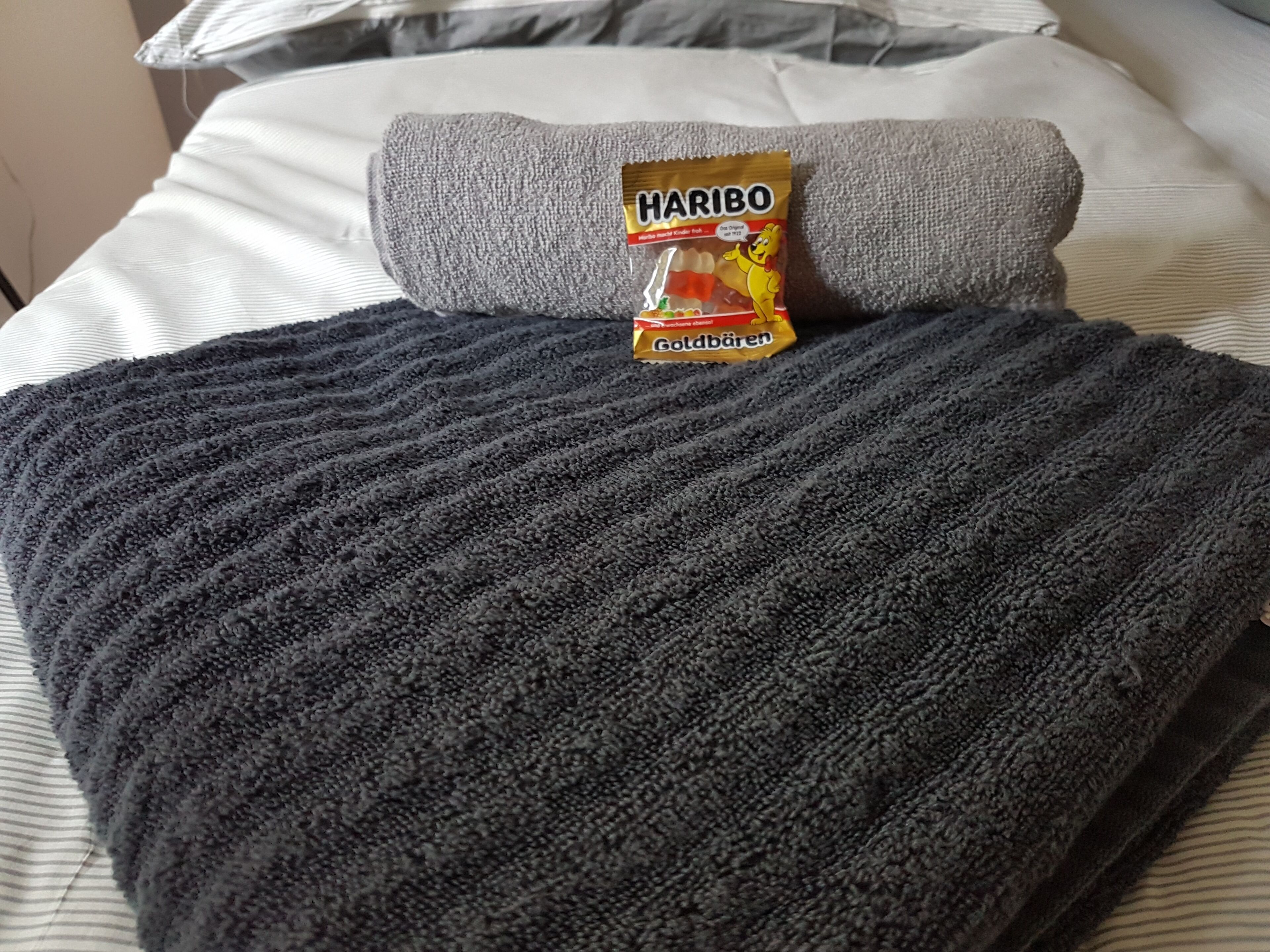 Ein Bett mit grauer Decke, Kissen und einer Packung Haribo Goldbären.
